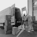 Dois trabalhadores usando equipamentos de proteção manuseiam encomendas junto a um veículo de entrega.