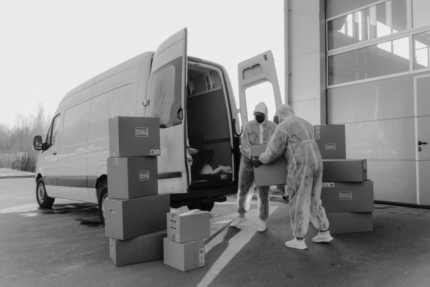 Dois trabalhadores usando equipamentos de proteção manuseiam encomendas junto a um veículo de entrega.