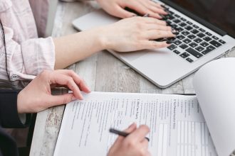 Mãos digitam em um laptop enquanto revisam documentos sobre uma mesa.