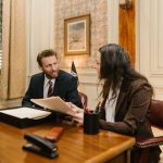 Dois profissionais conversam em um escritório enquanto analisam documentos sobre uma mesa.