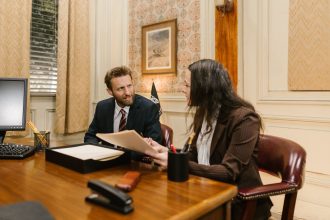 Dois profissionais conversam em um escritório enquanto analisam documentos sobre uma mesa.