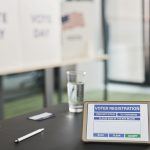 Tablet exibindo uma tela de registro eleitoral em uma estação de votação interna, com sinalização sobre o dia da votação.