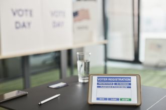 Tablet exibindo uma tela de registro eleitoral em uma estação de votação interna, com sinalização sobre o dia da votação.
