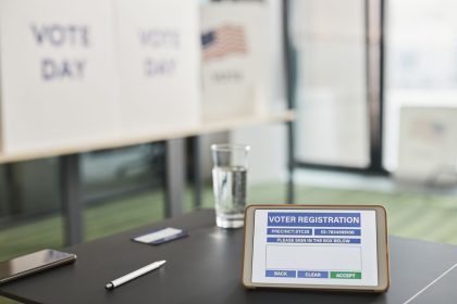 Tablet exibindo uma tela de registro eleitoral em uma estação de votação interna, com sinalização sobre o dia da votação.