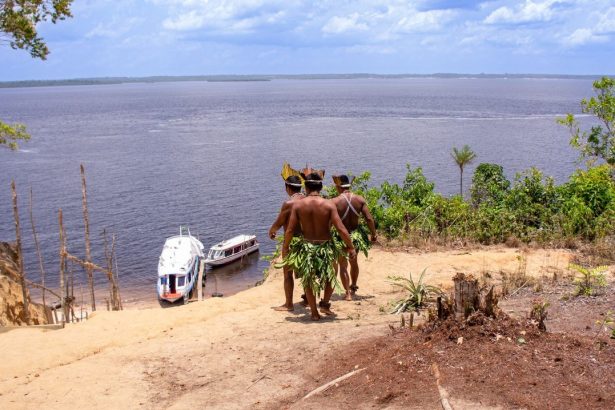 Dois homens indígenas observam barcos no rio Amazonas.