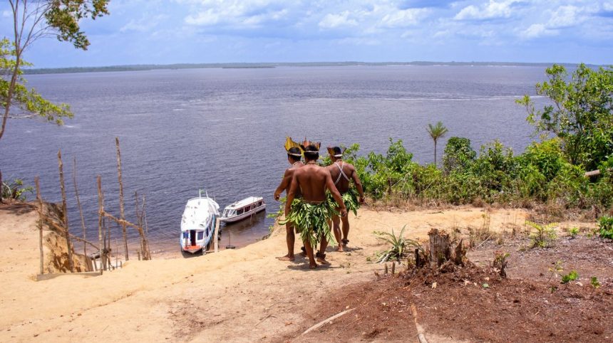 Dois homens indígenas observam barcos no rio Amazonas.
