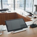 Sala de reunião institucional com mesa de madeira, microfones e telas digitais ao fundo.