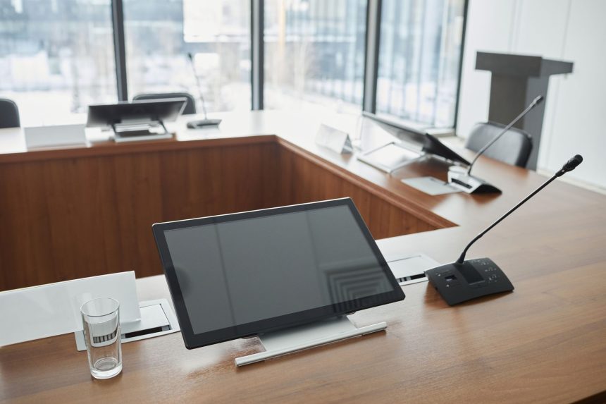 Sala de reunião institucional com mesa de madeira, microfones e telas digitais ao fundo.